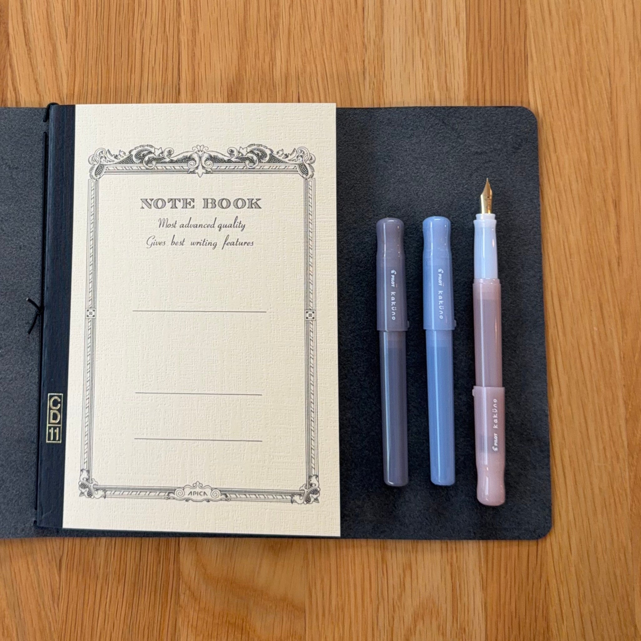 Open A5 Journey cover in Black with an Apica CD Notebook in Yellow, with three Kakuno Madoromi fountain pens. The two capped pens in Gray and Periwinkle, and one uncapped in light pink.
