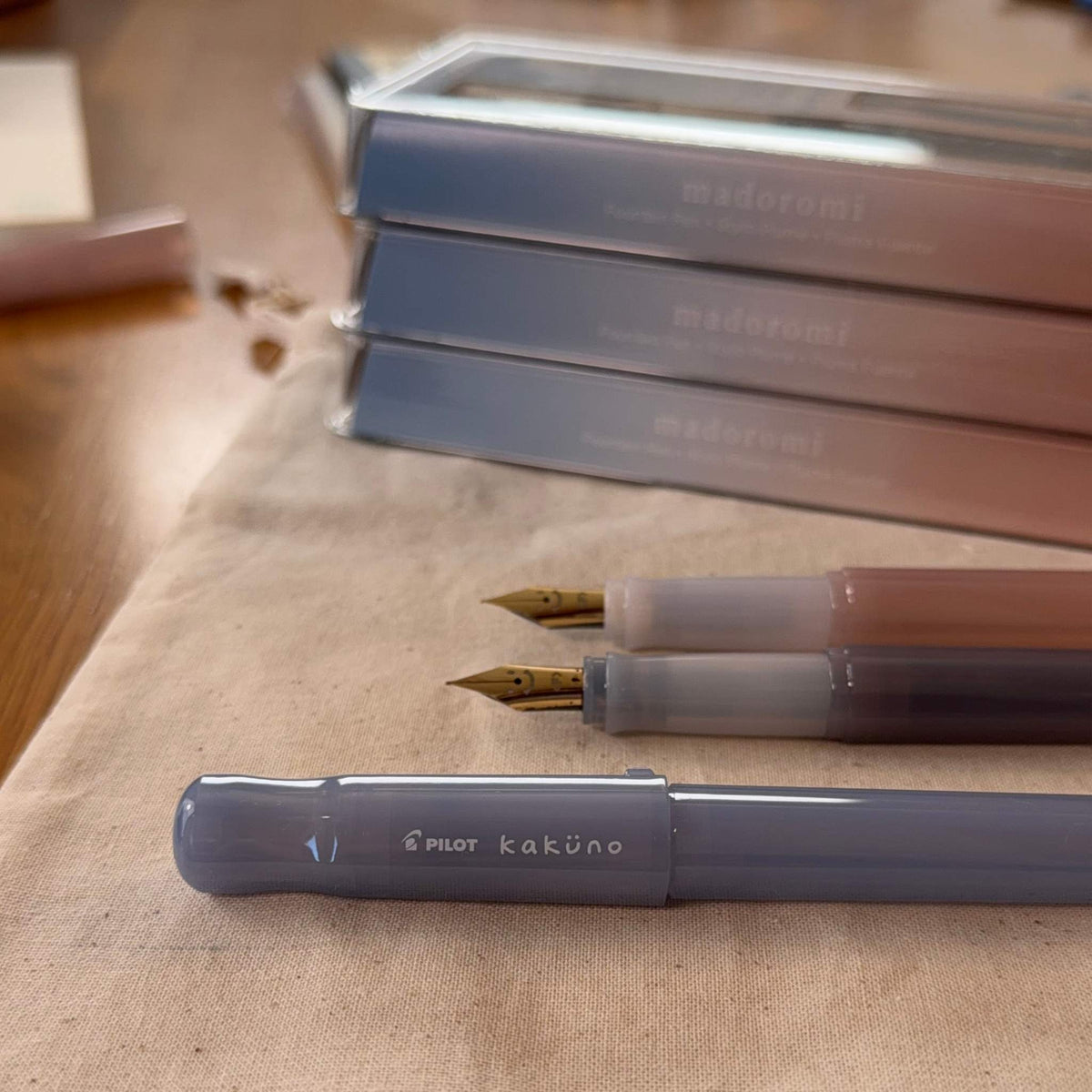 Pilot Kakuno Madoromi fountain pens in Periwinkle, Gray, and Light Pink, with the Gray and Light Pink pens uncapped to show the smiley face nib detail, and boxed Madoromi pens in the background
