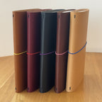 Five Journey A5 leather notebook covers in Cognac, Bordeaux, Black, Tobacco, and Natural, standing side by side