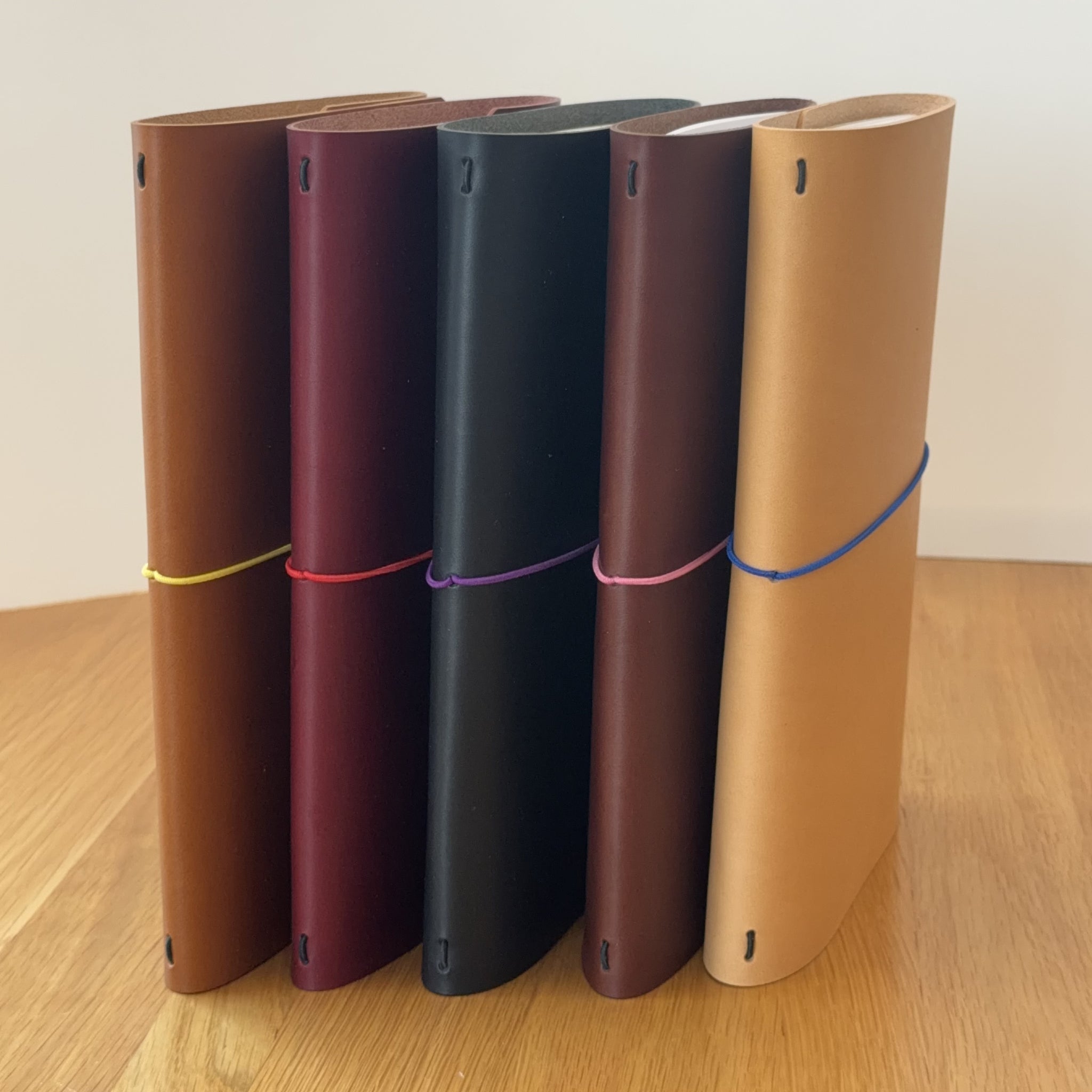 Five Journey A5 leather notebook covers in Cognac, Bordeaux, Black, Tobacco, and Natural, standing side by side