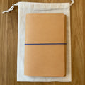 Journey A5 leather notebook cover in Natural, closed, resting on its Floramoon cotton dust bag