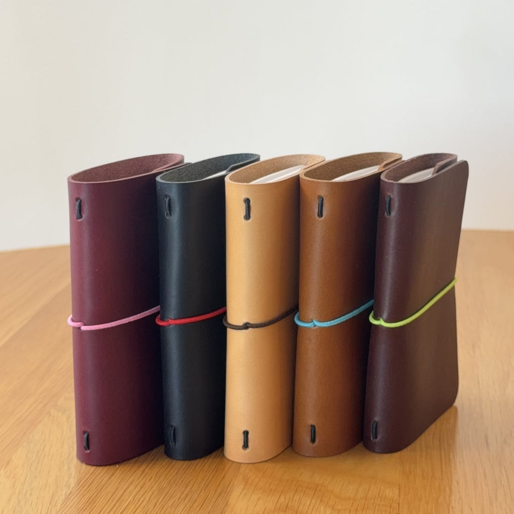 Five Journey A7 leather notebook covers in Bordeaux, Black, Natural, Cognac, and Tobacco, standing side by side
