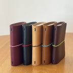 Five Journey A7 leather notebook covers in Bordeaux, Black, Natural, Cognac, and Tobacco, standing side by side