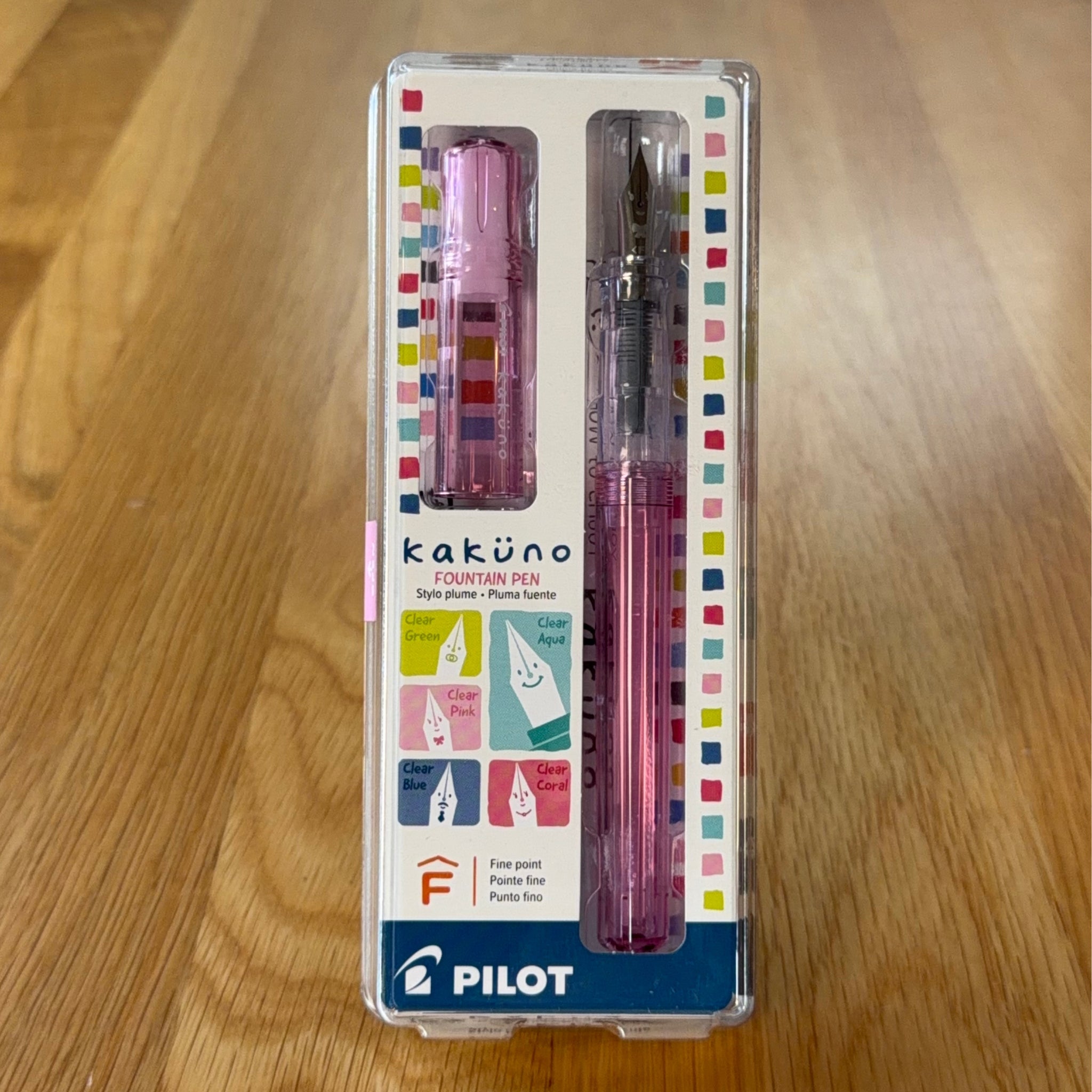 Pilot Kakuno Fountain Pen in Clear Pink, shown in its original packaging on a wooden surface, with the translucent barrel and stainless steel nib visible through the case