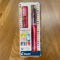 Pilot Kakuno Fountain Pen in Clear Coral, shown in its original packaging on a wooden surface, with the translucent barrel and stainless steel nib visible through the case