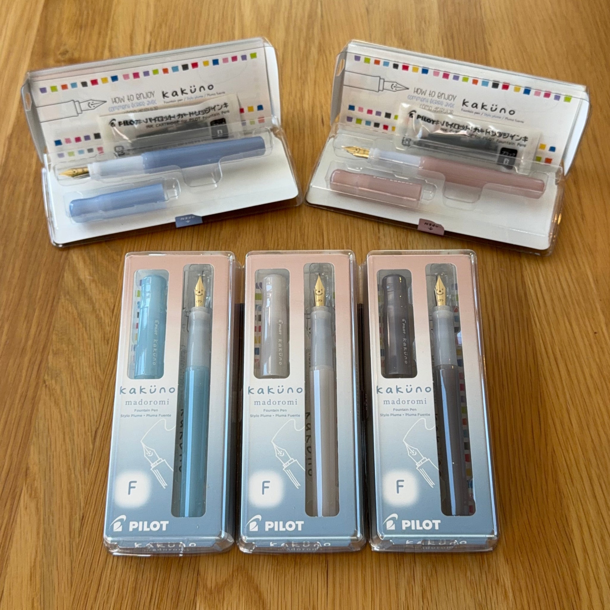 Five Pilot Kakuno fountain pens in their original packaging, showing the full range of colors (Light Blue, Light Pink, Periwinkle, White, Gray) arranged on a wooden table, with two packs shown open to display the pen bodies and ink cartridges.