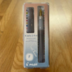 Pilot Kakuno Madoromi Fountain Pen in Gray, shown in its original packaging on a wooden surface, with the translucent barrel and gold nib visible through the case