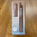 Pilot Kakuno Madoromi Fountain Pen in Light Pink, shown in its original packaging on a wooden surface, with the translucent barrel and gold nib visible through the case