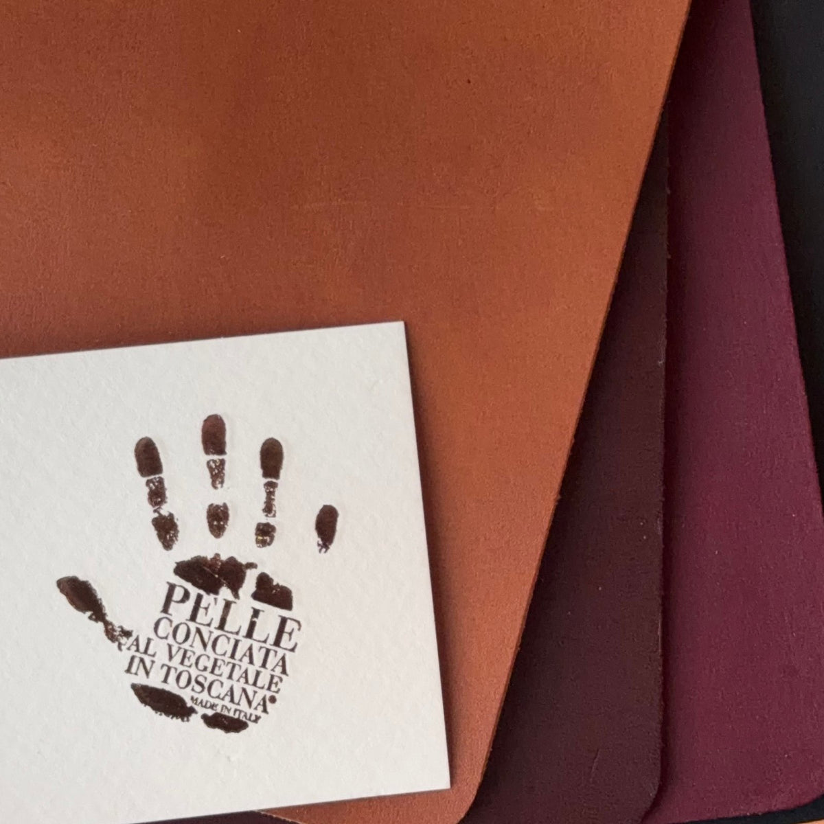 A close-up of a white square card stamped with a dark brown handprint and the text "PELLE CONCIATA AL VEGETALE IN TOSCANA," layered over several textured leather sheets in Cognac, Tobacco, Bordeaux, and Black.
